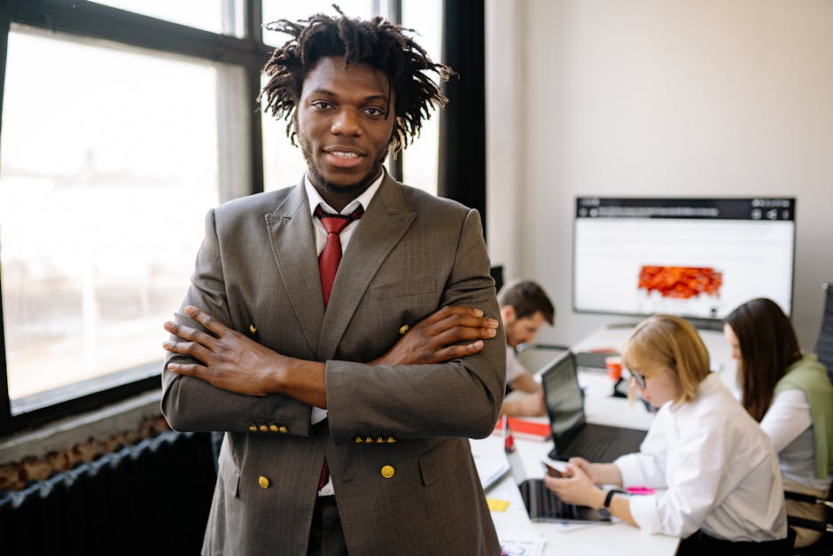 Confident businessman with arms crossed in a bright office environment with colleagues working in the background